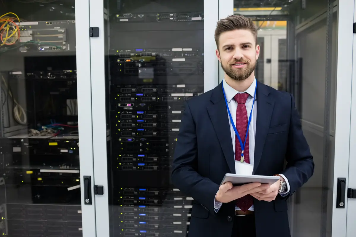 A man in a suit holding a tablet stands in front of server racks in a data center, overseeing AI and data strategy operations.